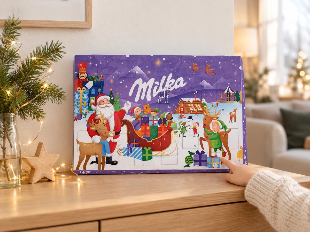 Milka adventní kalendář 200 g postavený na komodě s vánoční výzdobou, dětská ruka ukazuje na okénko